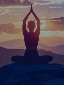A woman sitting in lotus pose with hands in prayer position above her head, by a lakeside.